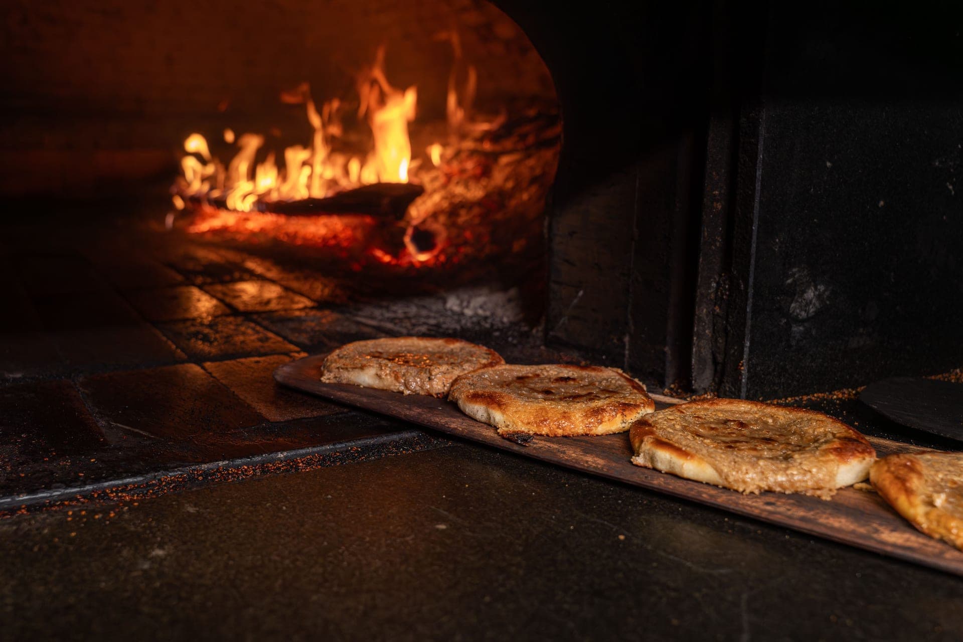 Flatbreads on a peel beside a wood-fired oven floor scattered with burnt flour