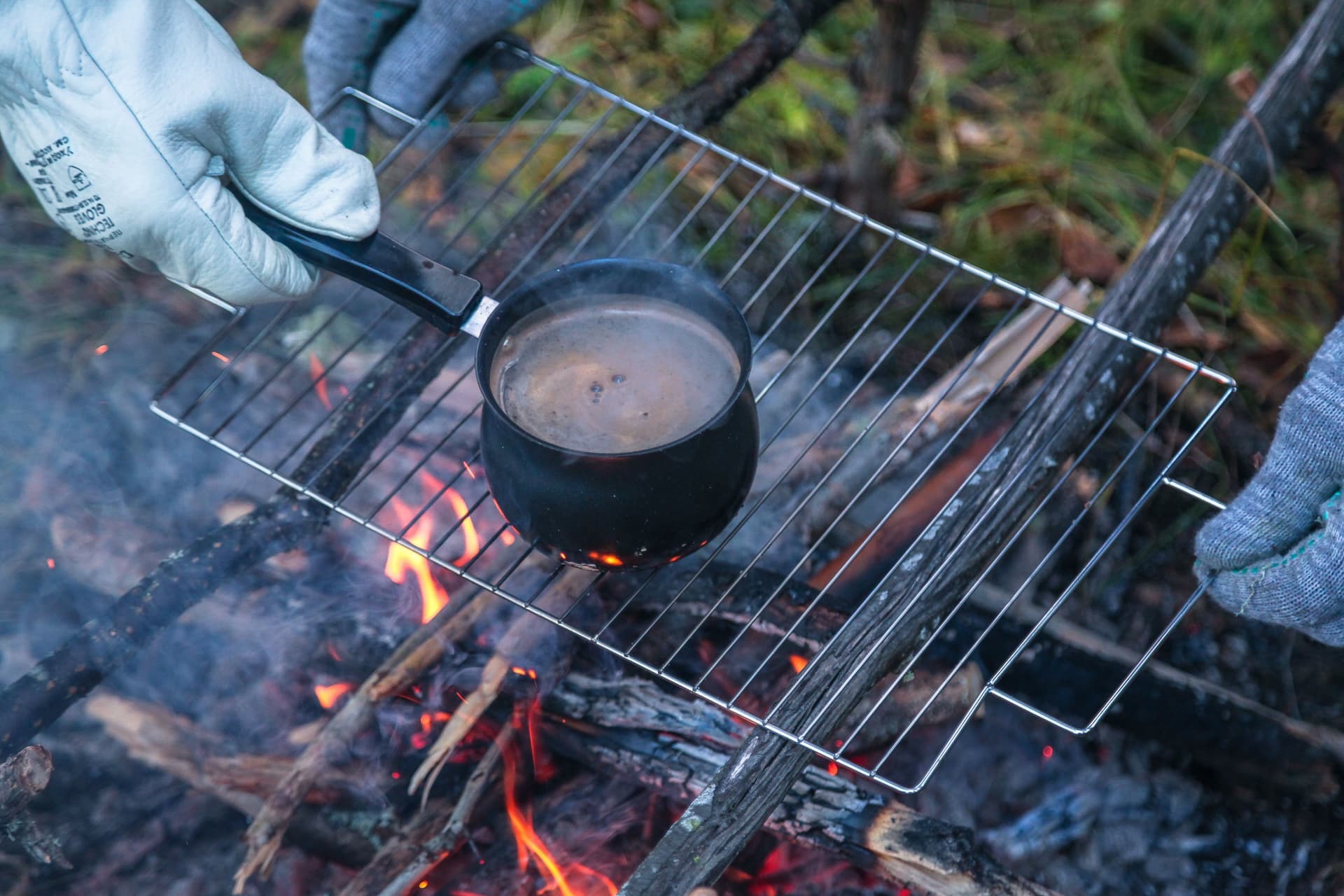 A leather-gloved hand lifting a hot pan from a grate over an open fire